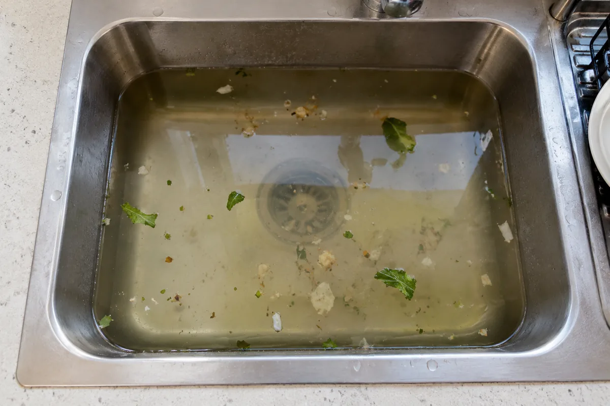 Fregadero de acero inoxidable atascado, con el agua estancada y restos de comida flotando